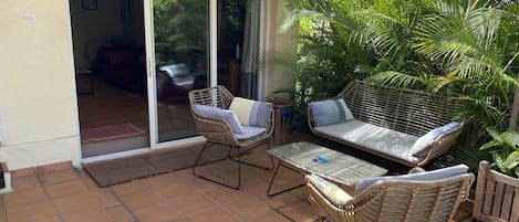Une terrasse dotée de meubles en osier, une porte vitrée donnant sur une pièce intérieure et une végétation luxuriante.