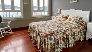 Un dormitorio con colcha estampada, una mecedora y vistas al agua a través de amplias ventanas.