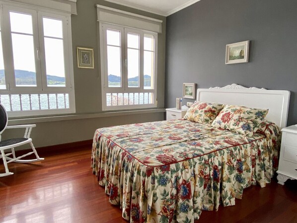 Un dormitorio con colcha estampada, una mecedora y vistas al agua a través de amplias ventanas.