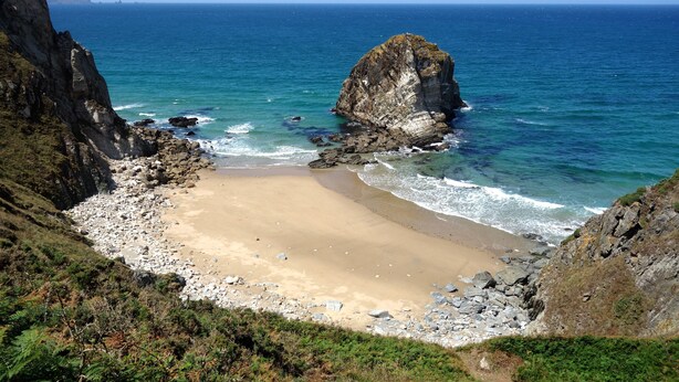 Un paisaje costero con playa rocosa, mar azul cristalino y una destacada formación rocosa.