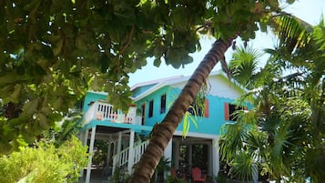 Una casa de dos pisos con un porche y una pequeña terraza, rodeada de vegetación tropical.
