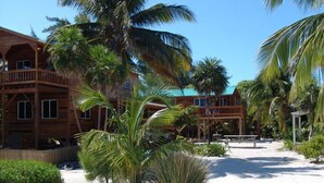 A wooden building with multiple balconies, surrounded by palm trees and a sandy area. - Caye Caulker Vacation Homes (Caye Caulker)