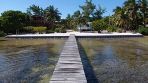 A wooden dock leading to a beach area with palm trees and a building in the background. - Caye Caulker Vacation Homes (Caye Caulker)