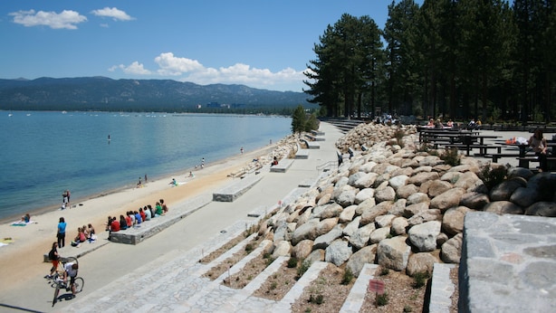 Une plage avec un escalier en pierre descendant jusqu’à l’eau, un espace aménagé pour pique-niquer et une rangée d’arbres.