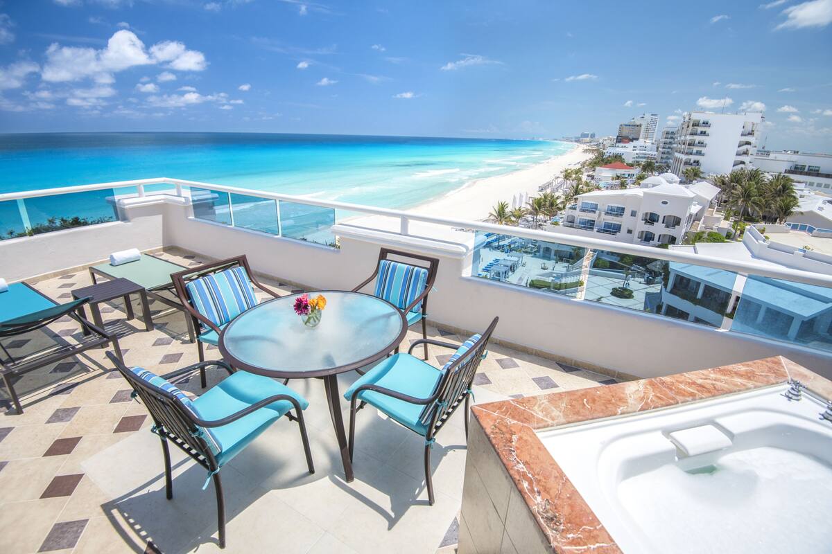A balcony with a glass table, chairs, and a hot tub overlooking a beach and resort buildings.