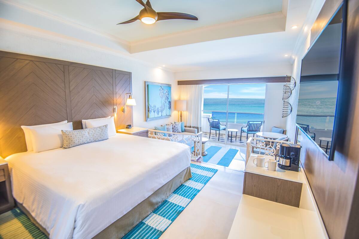 A hotel room with a large bed, a balcony with a view of the ocean, a dining area, and a ceiling fan.