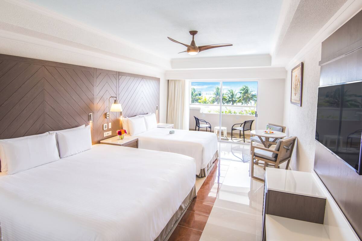 A hotel room with two beds, a ceiling fan, a TV, a balcony with a view of palm trees, and a dining area with chairs and a table.