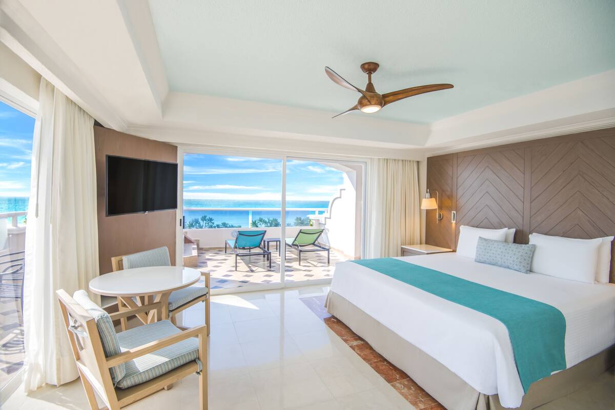 A hotel room with a large bed, a ceiling fan, a TV, a balcony with chairs, and a view of the ocean.