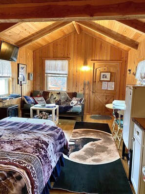 A cozy cabin with a wooden interior, a bed, a sofa, a dining table, and a refrigerator.
