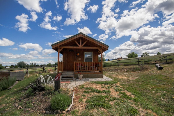 A wooden cabin with a porch, surrounded by a fenced field and a clear sky.