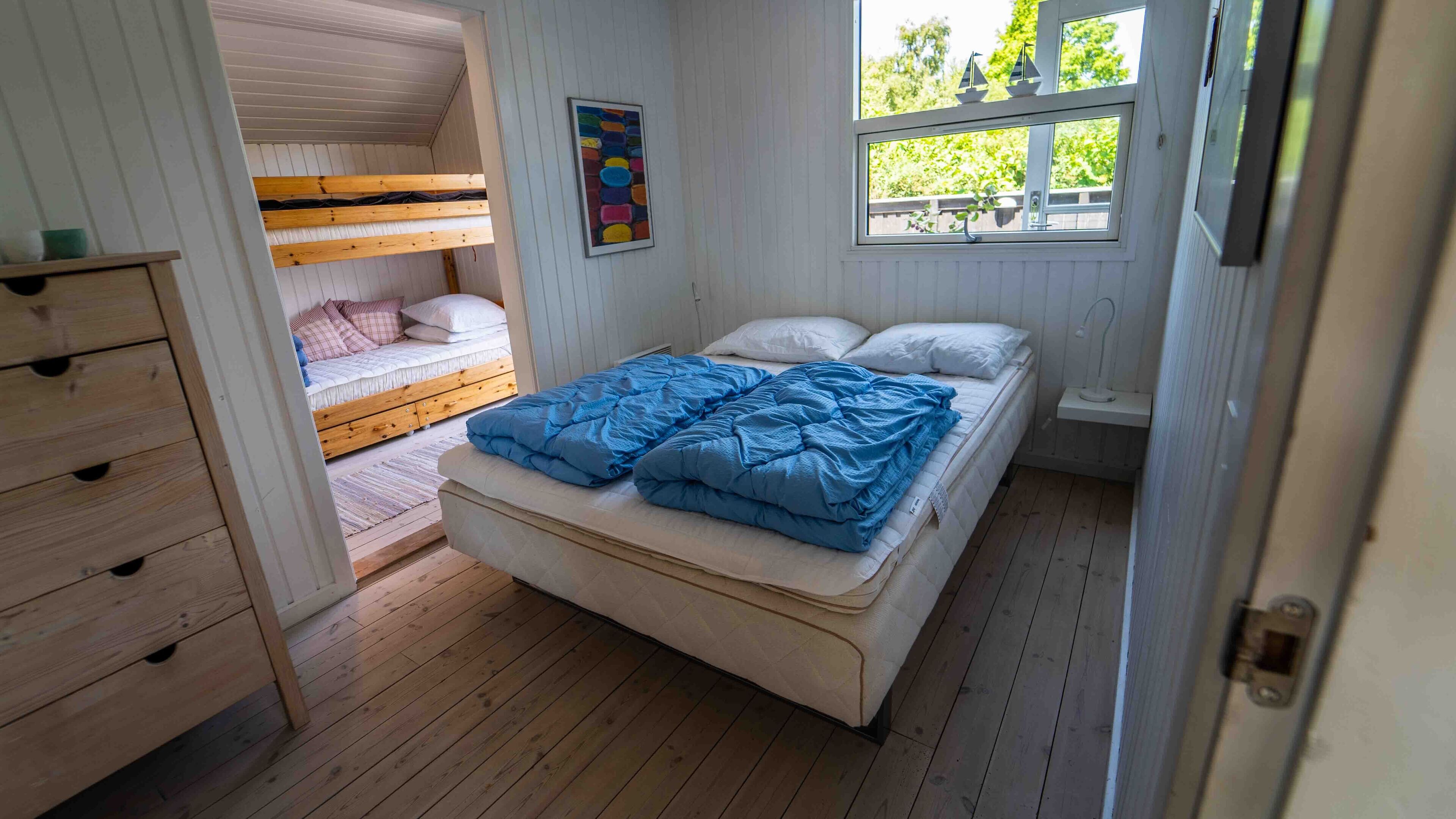 A room with a bunk bed, a single bed with blue bedding, a wooden dresser, and a window with a view of trees.