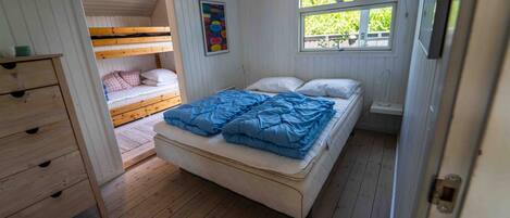 A room with a bunk bed, a single bed with blue bedding, a wooden dresser, and a window with a view of trees.