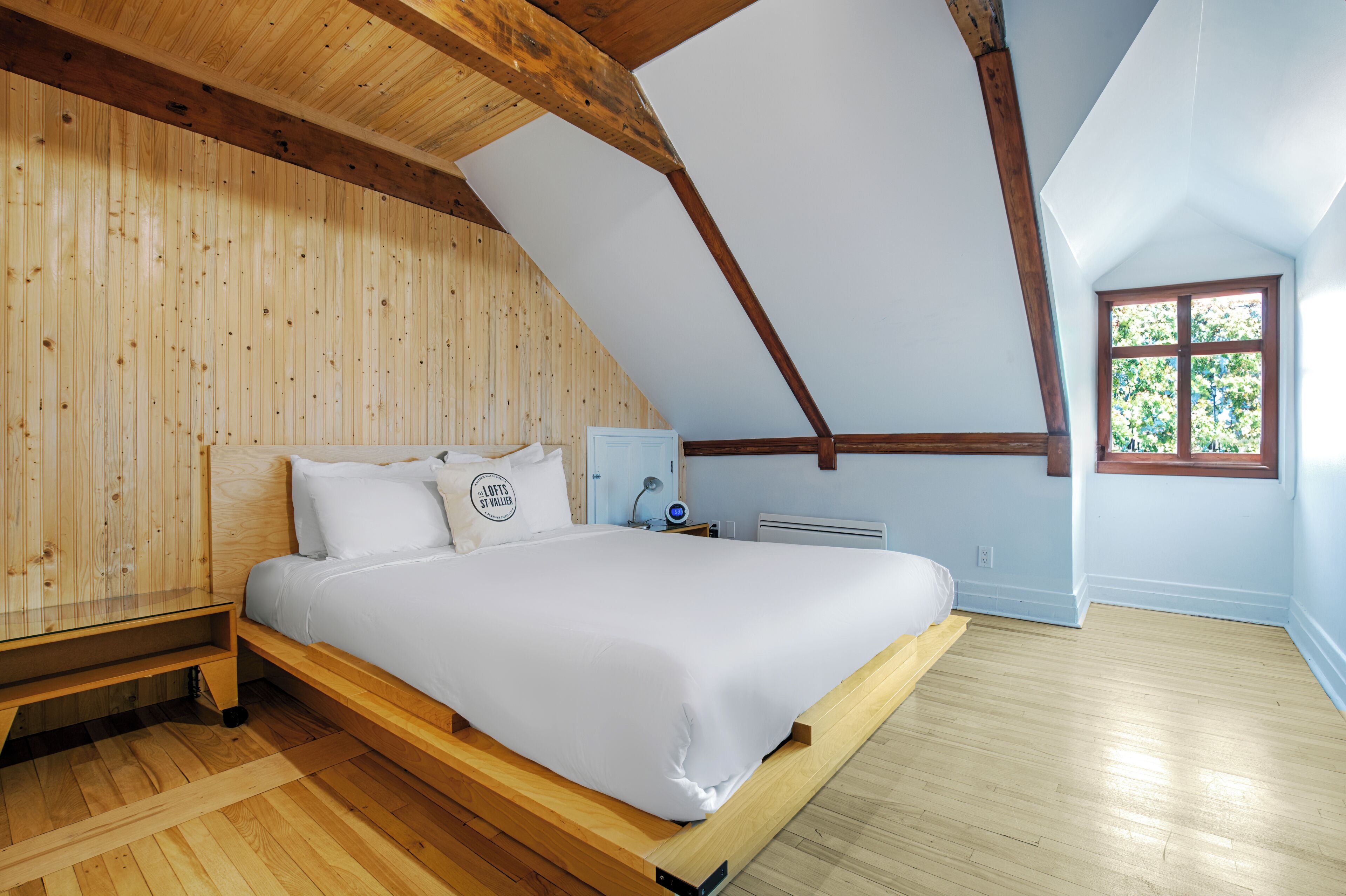 A bedroom with a sloped ceiling, wooden walls, a bed with white bedding, a bedside table, and a window with a view of trees.