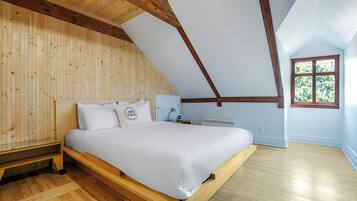 A bedroom with a sloped ceiling, wooden walls, a bed with white bedding, a bedside table, and a window with a view of trees.
