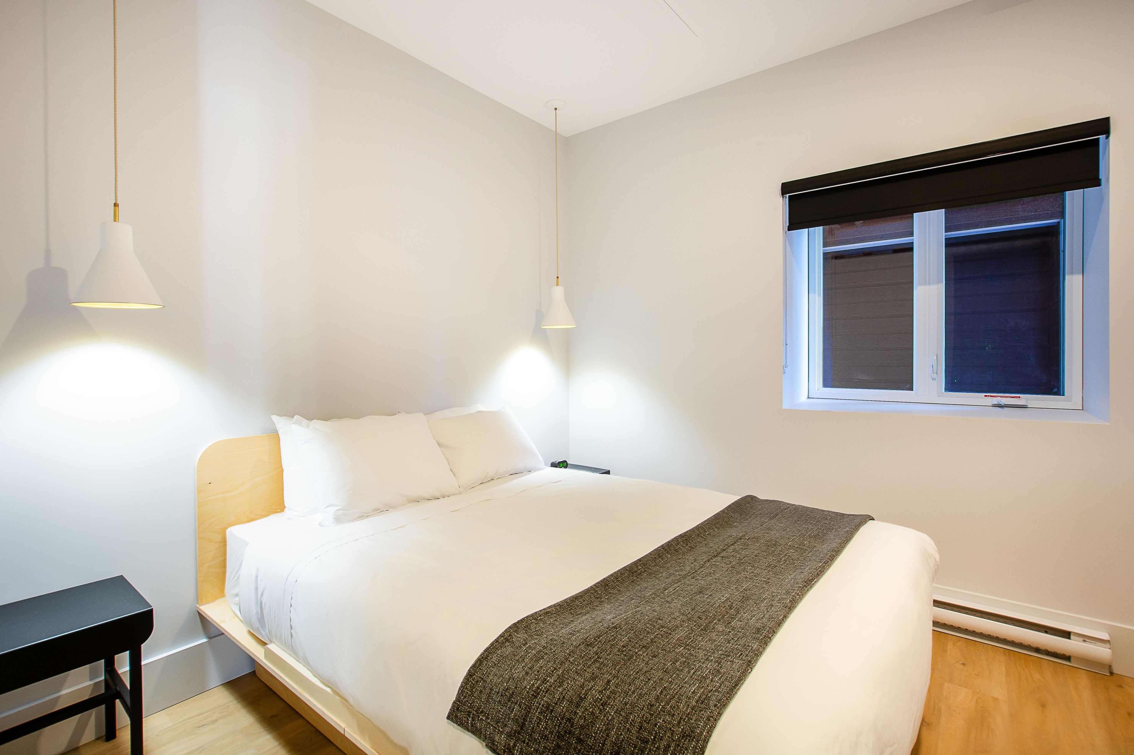Una cama bien hecha con ropa de cama blanca y una manta gris, una mesita de noche y una ventana con persiana negra.