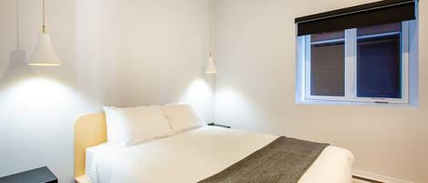 Una cama bien hecha con ropa de cama blanca y una manta gris, una mesita de noche y una ventana con persiana negra.