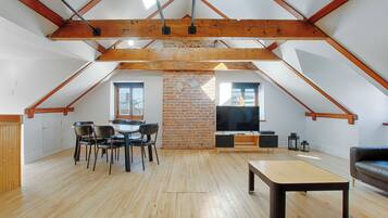 An attic-style room with exposed wooden beams, a brick wall, a dining table with chairs, a flat-screen TV, and a sofa.