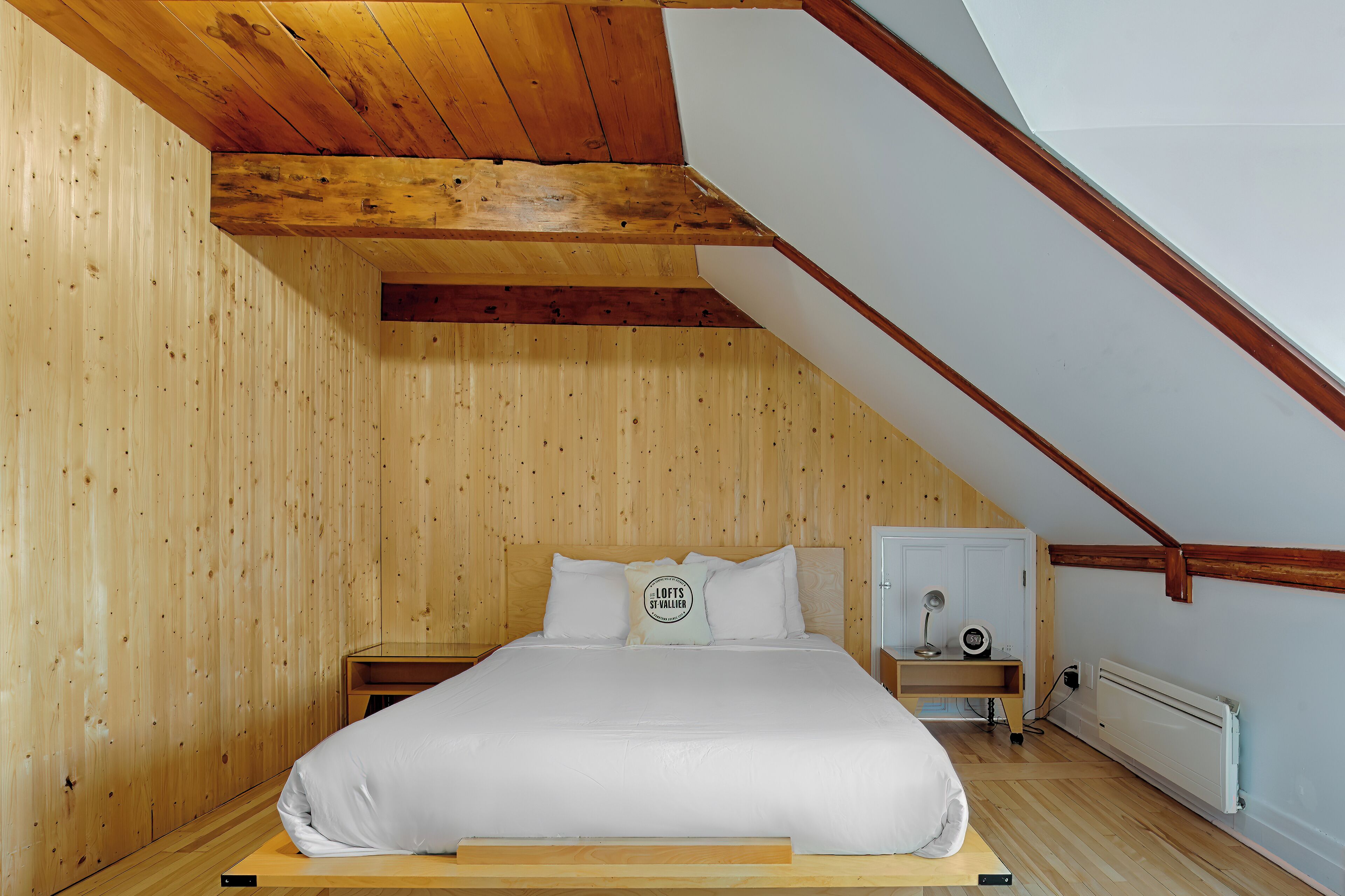 A bedroom with a sloped ceiling, wooden walls, a bed with white bedding, a nightstand, a desk with a lamp, and a radiator.