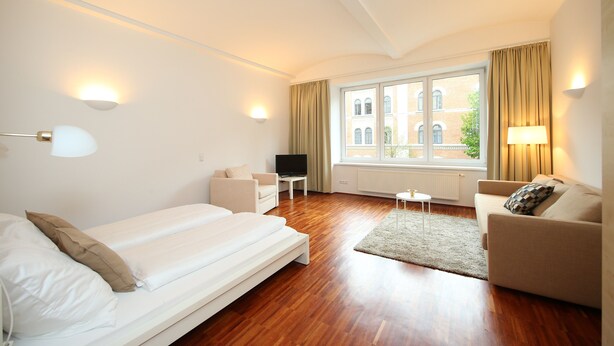 Ein modernes Hotelzimmer mit Bett, Sofa, Fernseher und Blick auf die Stadt durch große Fenster.