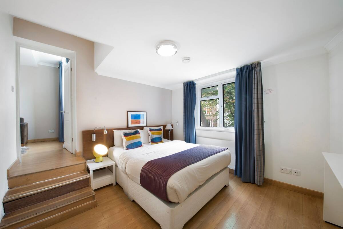 Une chambre à coucher avec un lit, une table de chevet, une lampe, une fenêtre avec des rideaux et une vue sur l’extérieur.