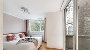 A bedroom with a large window, a bed with white bedding and red pillows, a bedside table with a lamp, and a view of a brick building outside. - Lux London South Kensington (London)