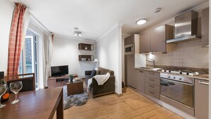 A modern kitchen with a built-in oven, microwave, and stainless steel cabinets. There is a dining area with a wooden table and chairs, and a cozy seating area with a sofa and a lamp. - Lux London South Kensington (London)