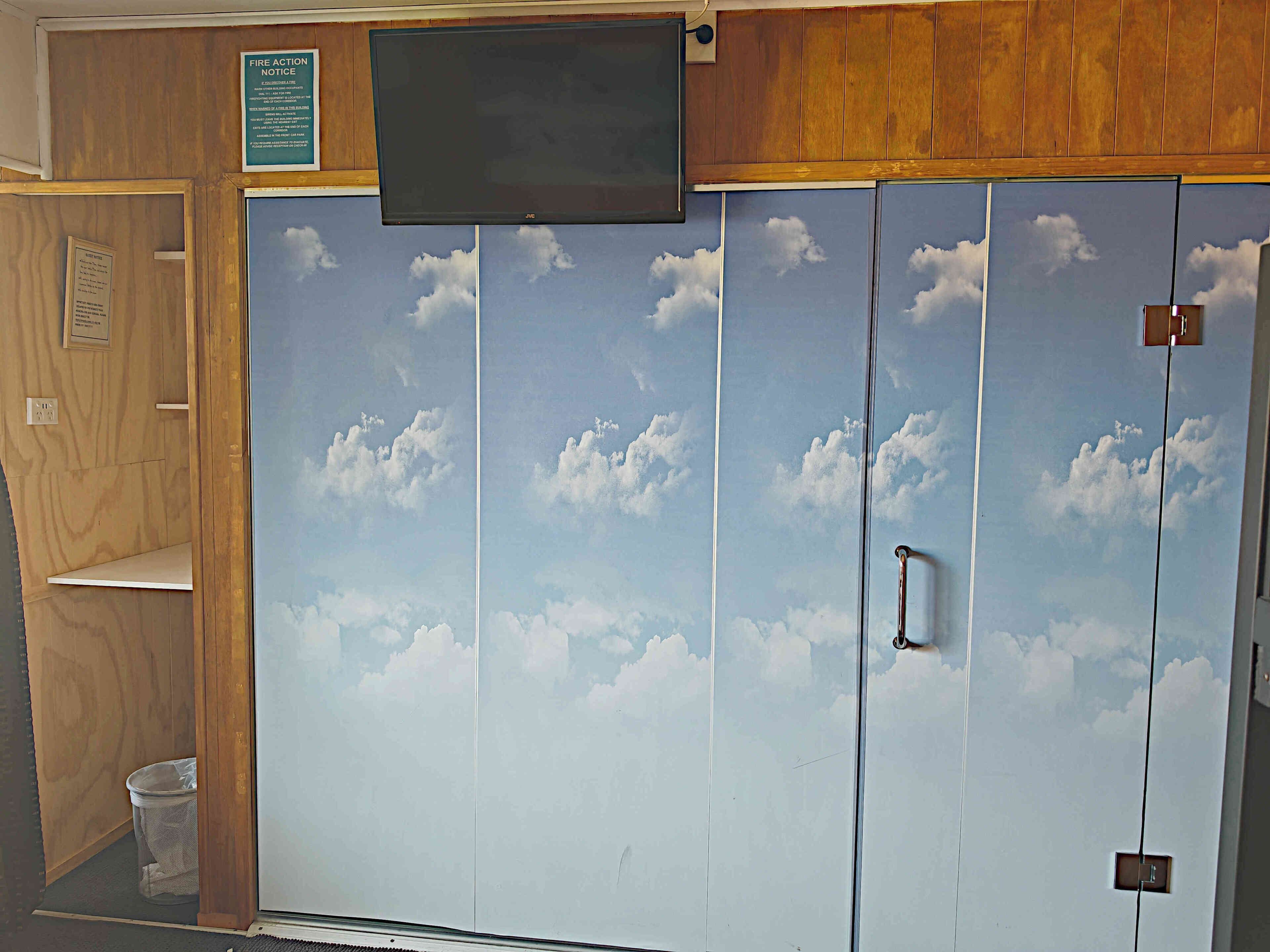A room with a large wardrobe featuring a sky and clouds design, a television mounted above it, and a small trash bin to the left.