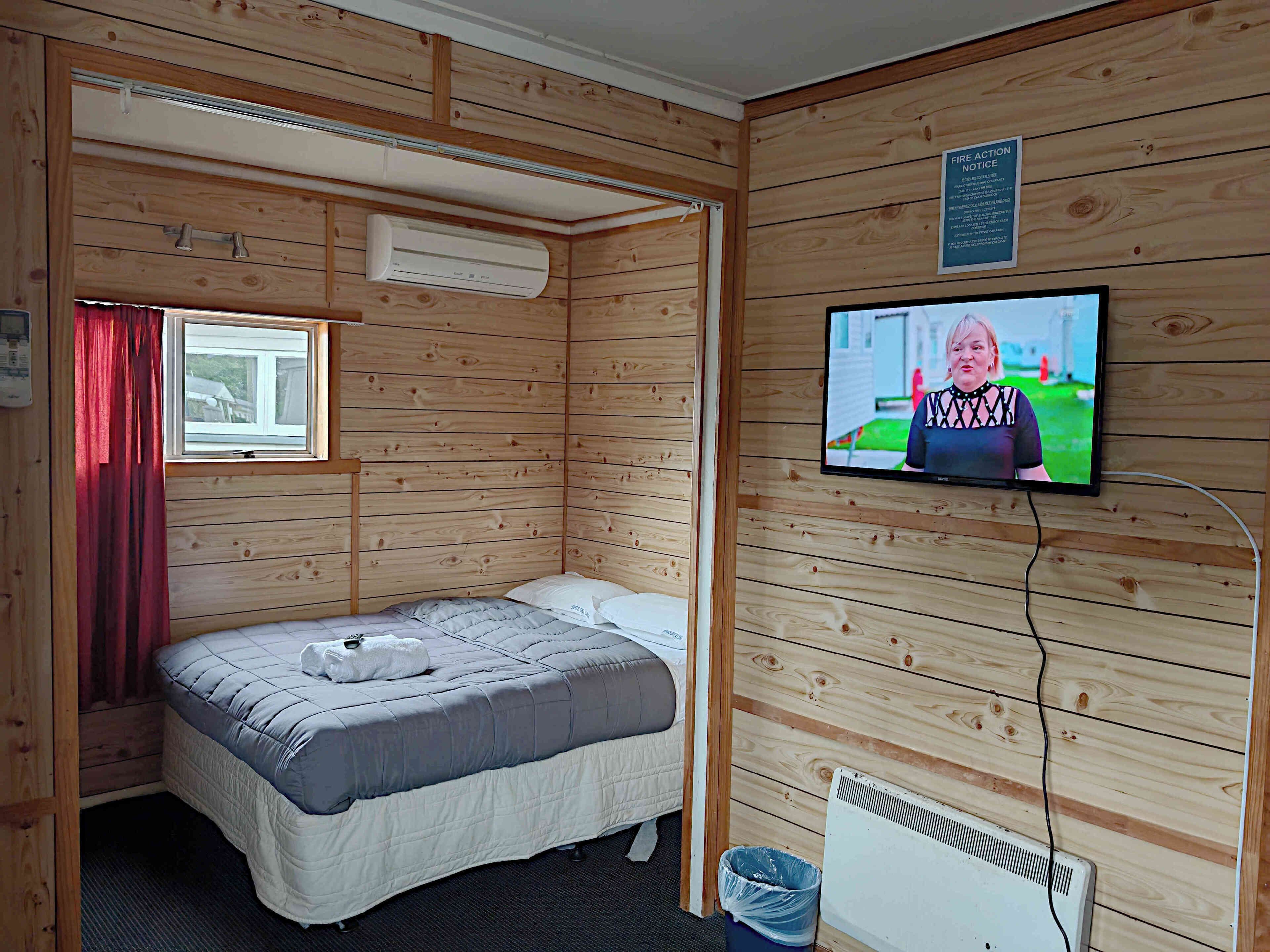 A wooden-paneled room with a bed, a window with red curtains, a wall-mounted TV, and a fire action notice.