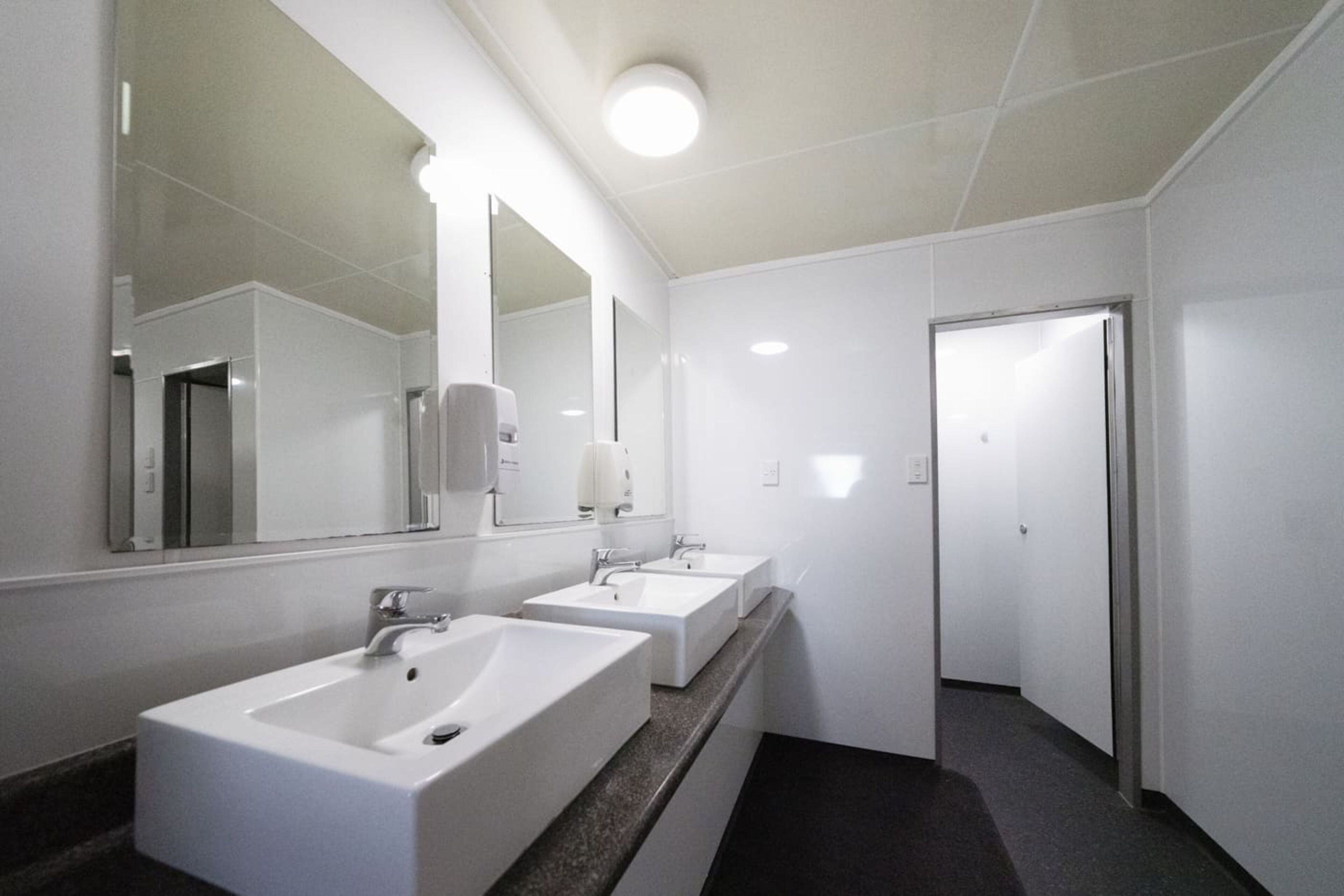 Un baño moderno y limpio con tres lavamanos, un espejo grande y una puerta que da a otra habitación.