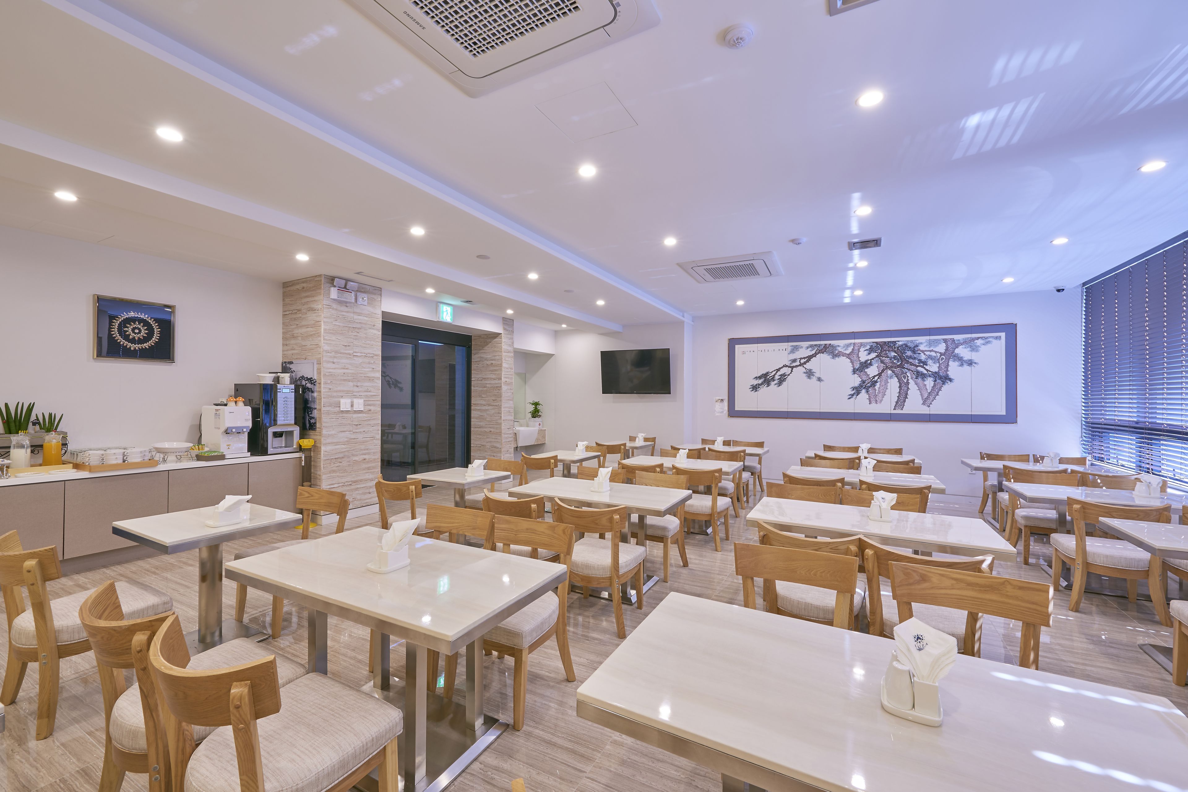 A spacious dining area with multiple tables and chairs, a kitchen counter with appliances, and a large framed artwork on the wall.