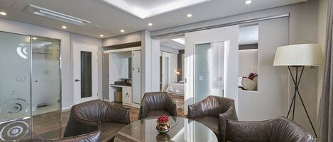 A modern hotel room with a round glass table, leather chairs, a lamp, and a view of the kitchen and bedroom.