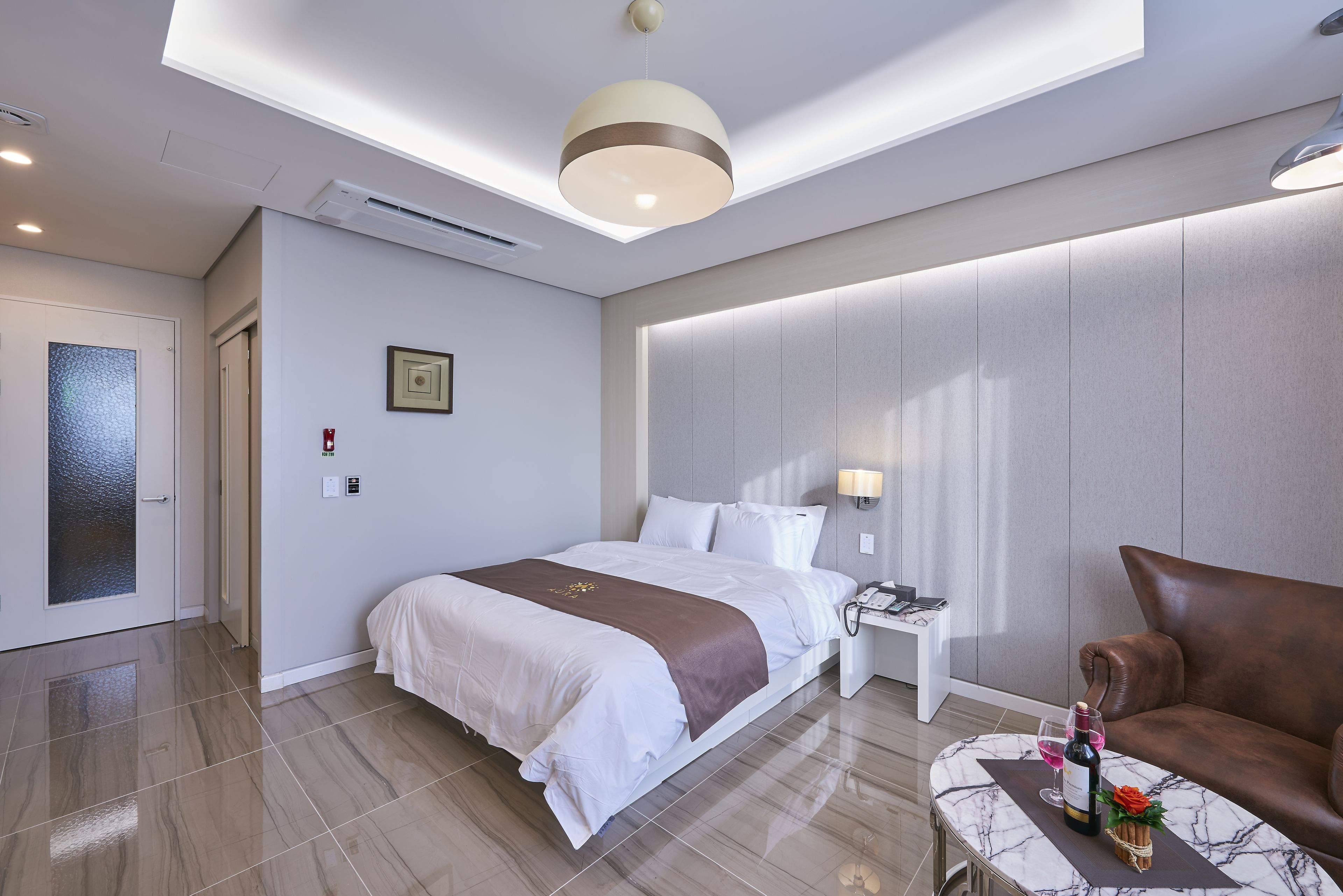 A modern hotel room with a large bed, a brown leather sofa, a marble coffee table, and a wall-mounted lamp.