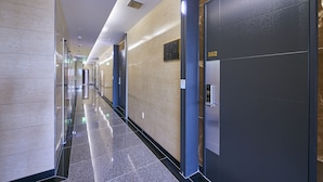 Un couloir d’hôtel moderne, avec un sol en marbre poli, un ascenseur en verre et une porte d’ascenseur sombre portant le numéro 602.