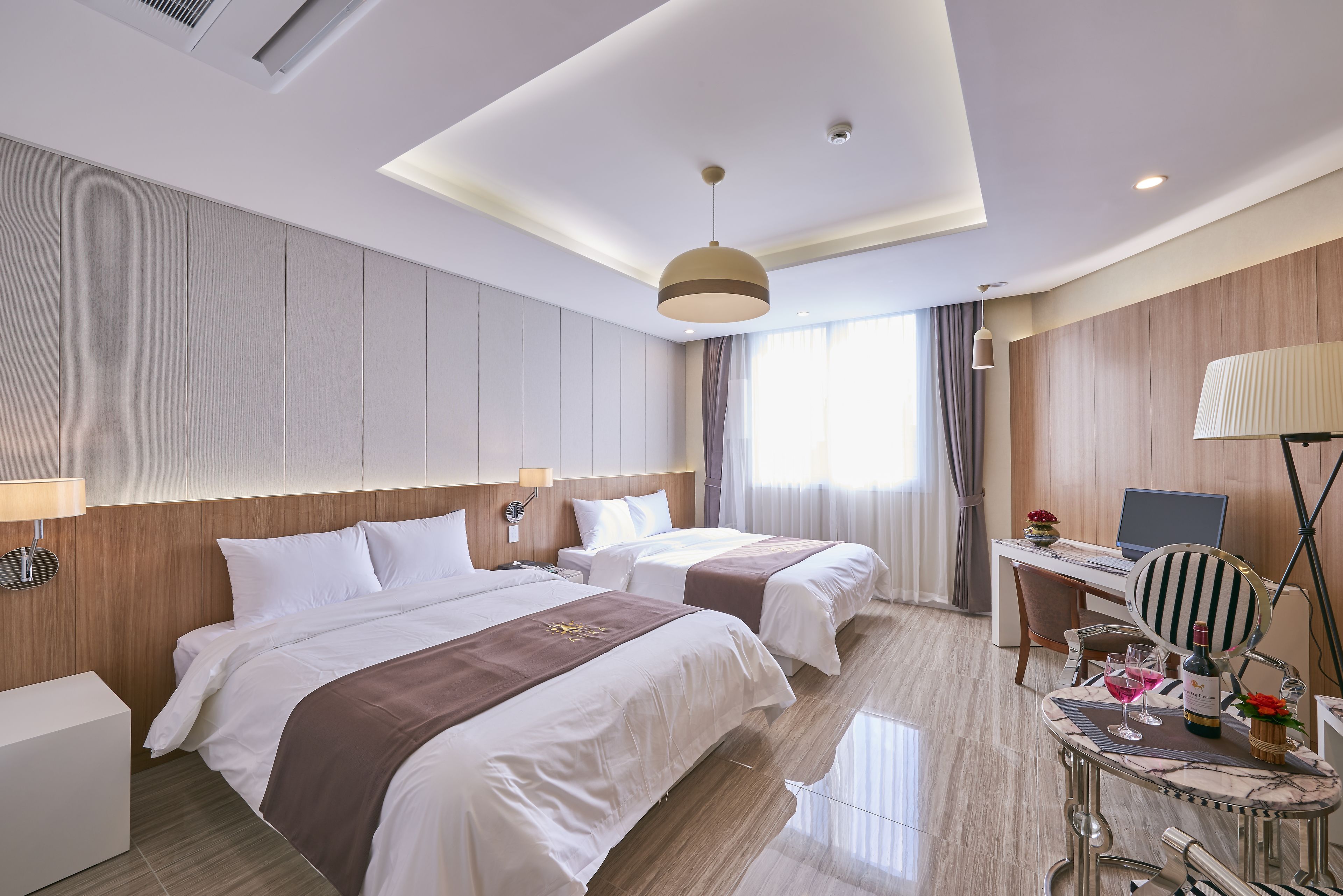 A hotel room with two beds, a desk with a computer, a chair, and a table with a wine bottle and glasses.
