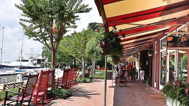 Un paseo marítimo con sillas rojas, una zona cubierta con un cartel que dice 'Beachside of Hilton House', y barcos amarrados al fondo.
