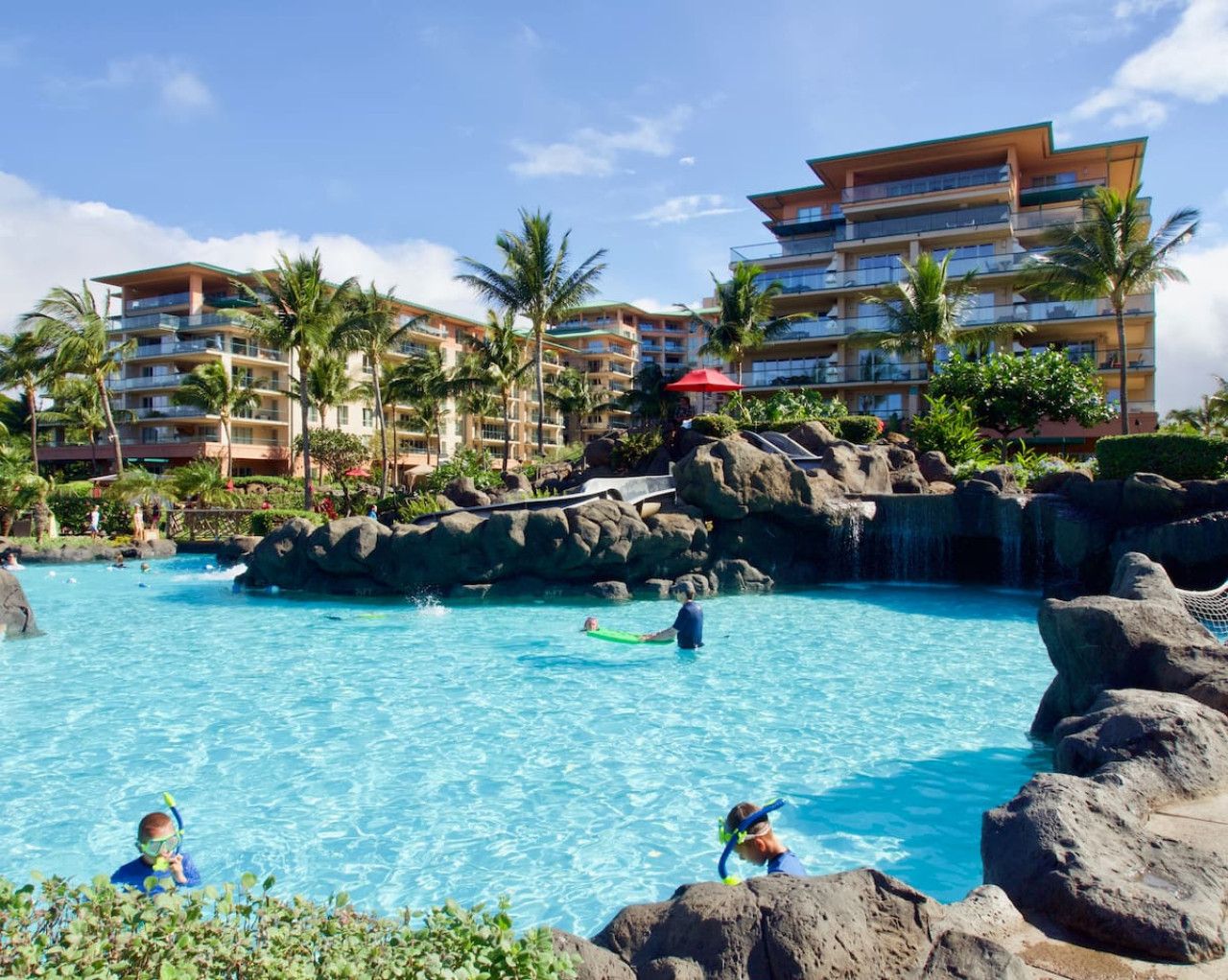 Zwembad van een resort met helderblauw water, omringd door palmbomen en rotsformaties.