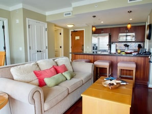 A living room with a sofa, a coffee table, and a kitchen area with a refrigerator, microwave, and bar stools. - Beautiful Ocean View on Quiet Side of Honua Kai (Lahaina)