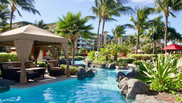 Ein Poolbereich eines Resorts mit Liegestühlen, einer überdachten Sitzgruppe und Palmen.