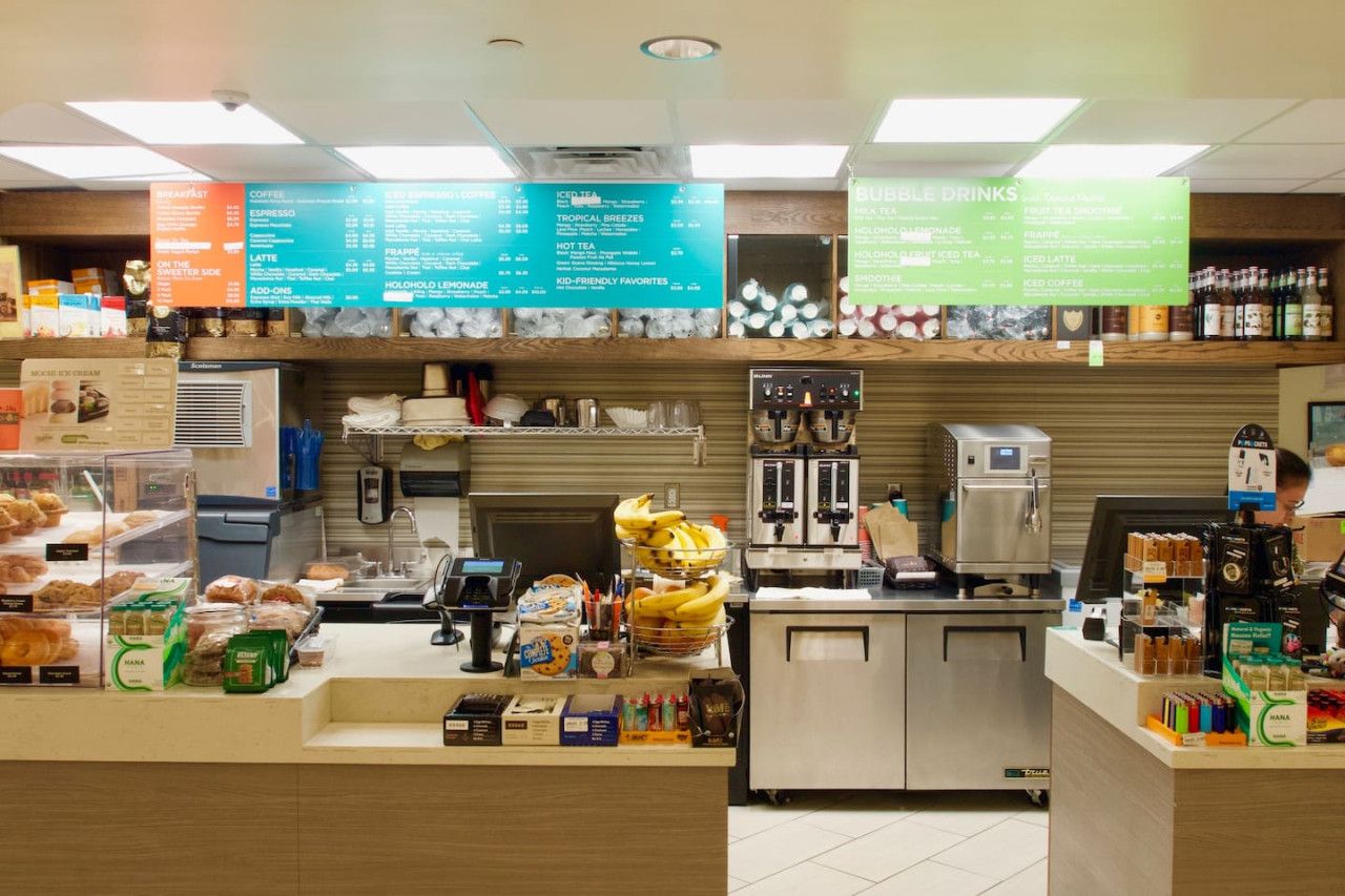 Interior de una cafetería con una pizarra de menú, máquinas de café y una variedad de productos horneados y bocadillos a la vista.