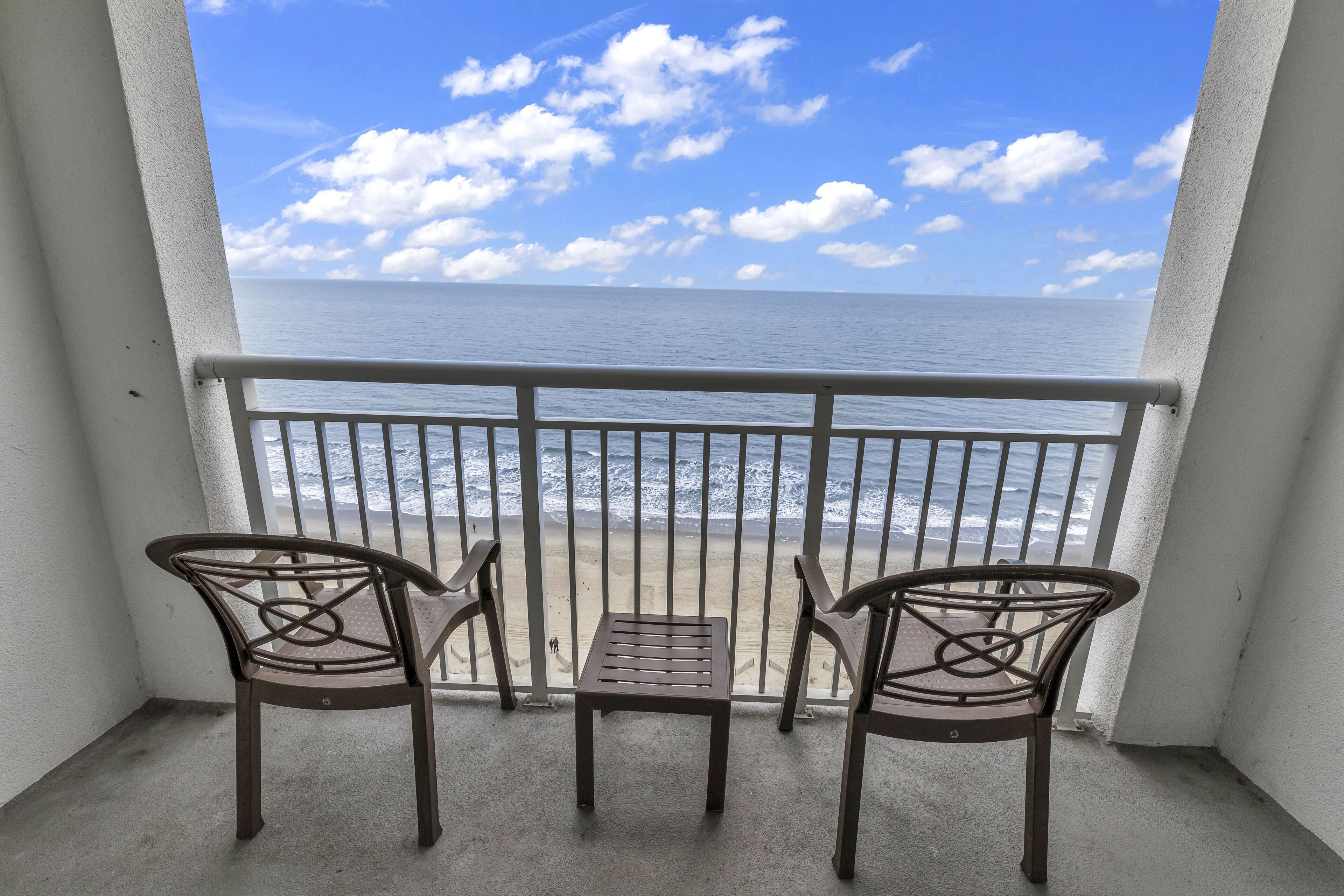 A balcony with two chairs and a small table, overlooking a beach with waves and a clear sky.