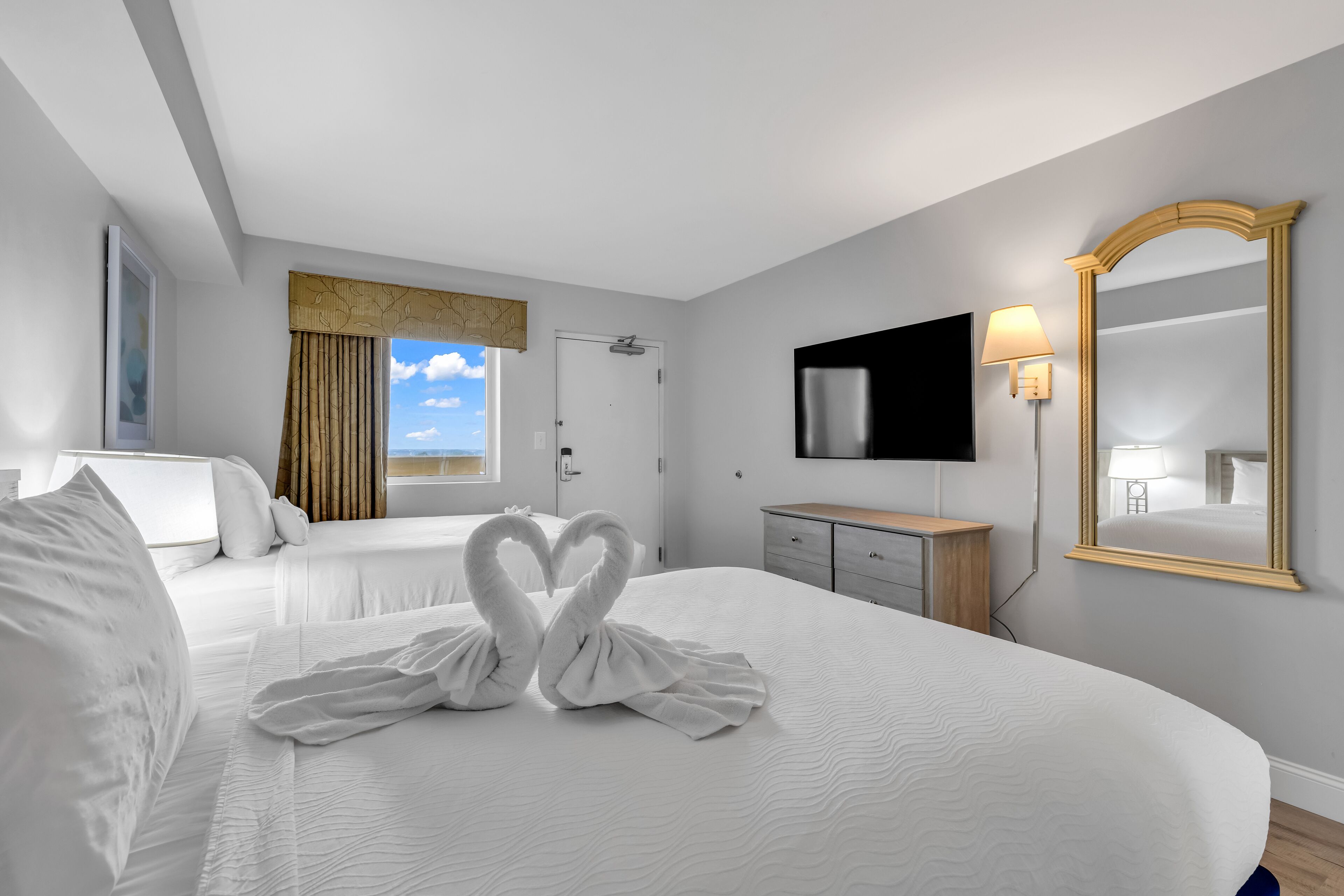 Una habitació d'hotel amb dos llits, televisió, mirall i finestra amb cortines.