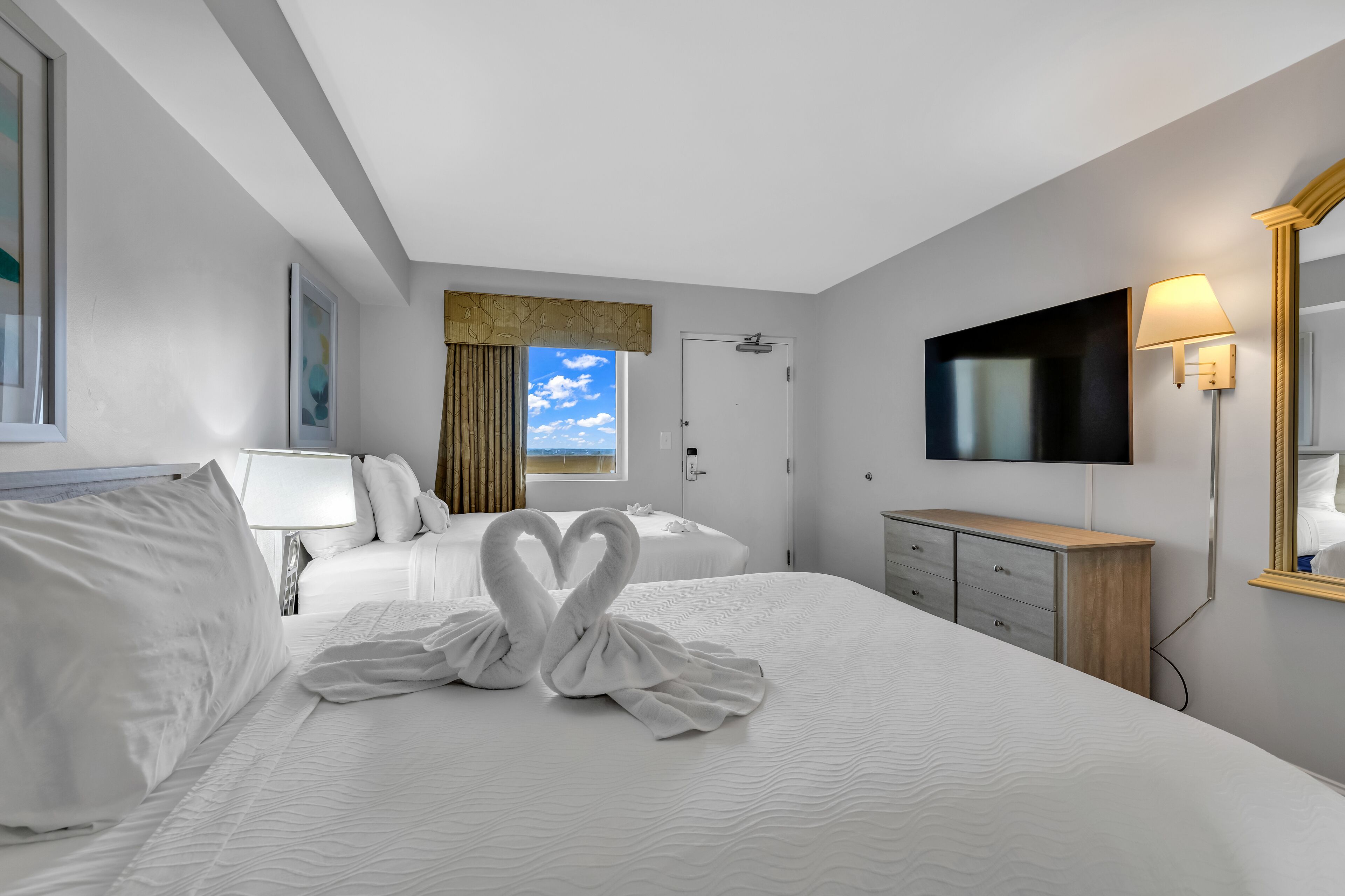 A hotel room with a large bed, two white pillows, a TV, a dresser, and a window with a view of the sky.