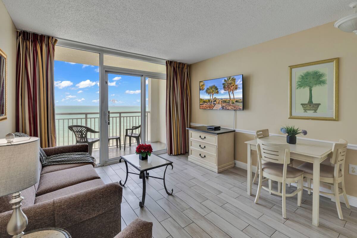 A balcony with a view of the ocean, a dining area with a table and chairs, a sofa, and a TV on the wall.