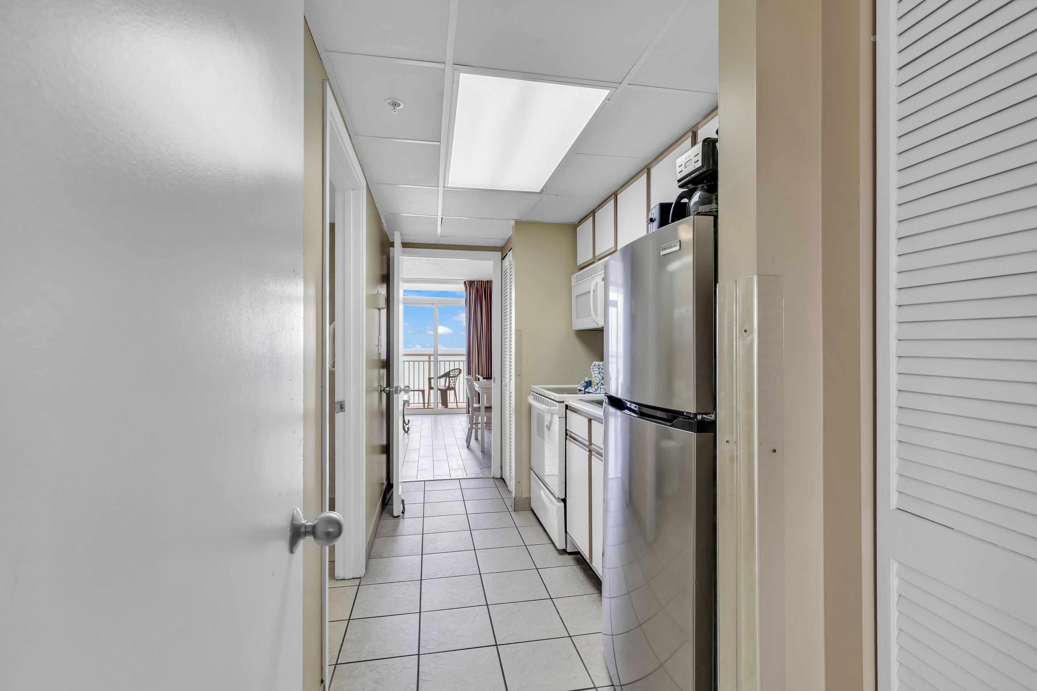 A narrow hallway with a white door on the left, a kitchen area with a refrigerator and microwave on the right, and a view of a balcony with chairs and a table.