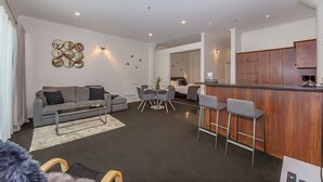 Een moderne woonkamer met een grijze bank, een salontafel, en een eethoek met tafel en stoelen. Er is een keuken met houten kastjes en een bargedeelte.
