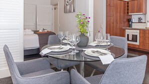 Een eetgedeelte met een ronde glazen tafel, gedekt voor vier personen, omringd door grijze fauteuils. Op tafel staat een vaas met paarse bloemen, en aan de muur hangt een schilderij.