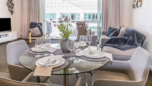 Een eetgedeelte met een glazen tafel gedekt voor vier personen, omringd door grijze fauteuils. Op tafel staat een vaas met bloemen, en er is een flatscreen televisie zichtbaar op de achtergrond.