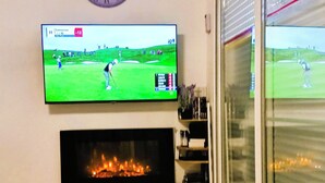 A living room with a flat-screen TV mounted on the wall displaying a golf game, a fireplace below the TV, and a glass door leading outside. - Antibes, Secure 1 bedroom apt, Sea Views, Swimming Pool, 3 adults - own parking (Antibes)