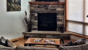 Una sala de estar con chimenea de piedra, un televisor de pantalla plana montado encima, un cuadro de un ciervo y una ventana con persianas.
