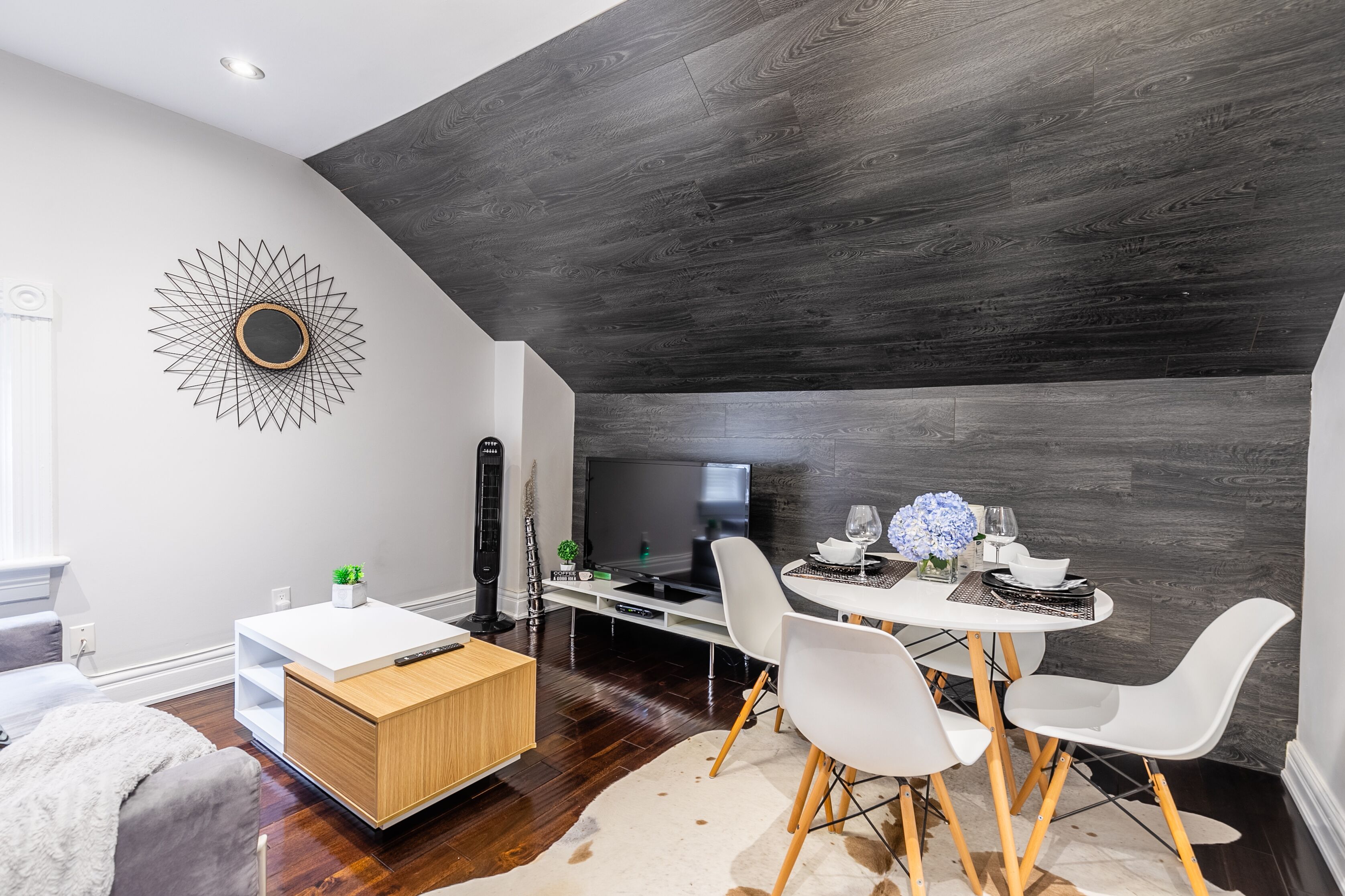 A modern living room with a dark wooden ceiling, a white dining table with four chairs, a white sofa, and a decorative wall piece.